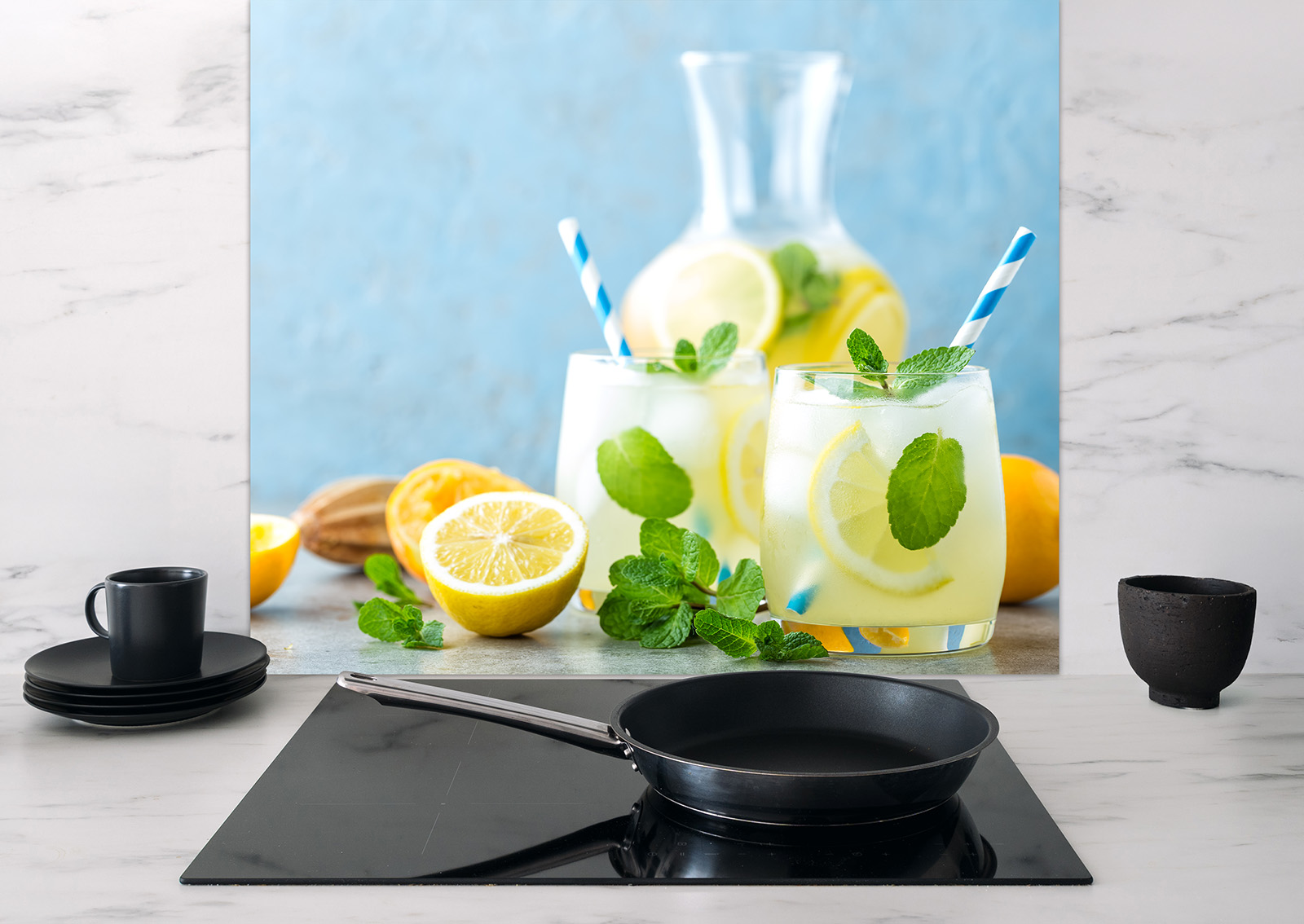 Spritzschutz mit Zitronenlimonade, Gläser mit Zitronenscheiben und Minze, Küchenutensilien im Vordergrund.